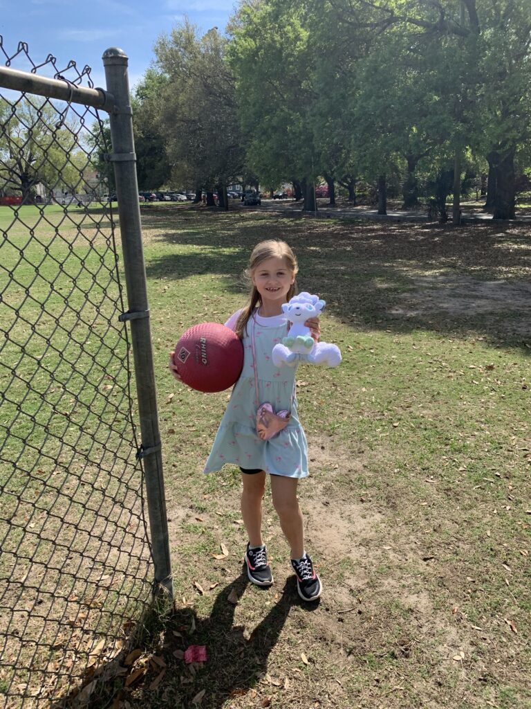 girl holds a kickball and a stuffed animal standing next to baseball fence